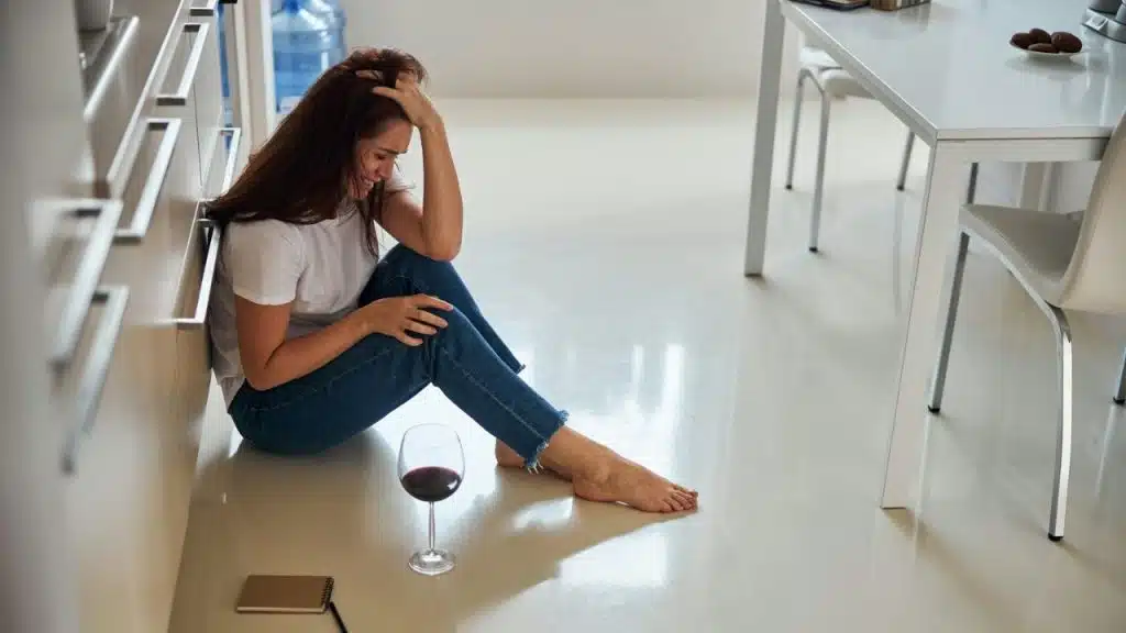 A woman sits on a kitchen floor, looking down with a glass of red wine nearby.