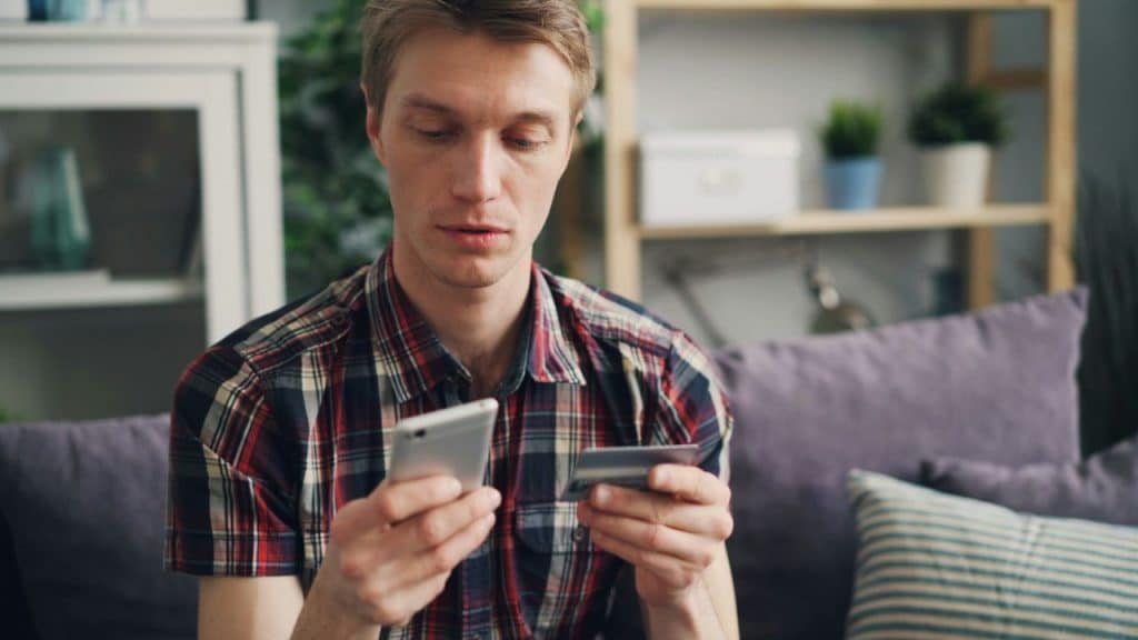 A man in a plaid shirt holds a credit card and a cell phone.