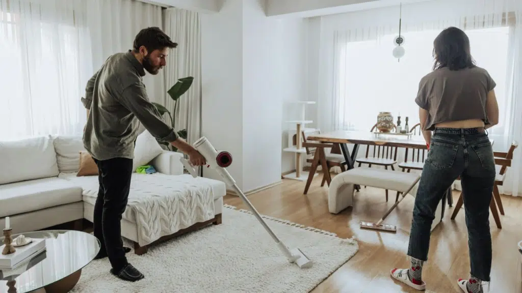A man vacuums a living room rug while a woman cleans the floor in the background.