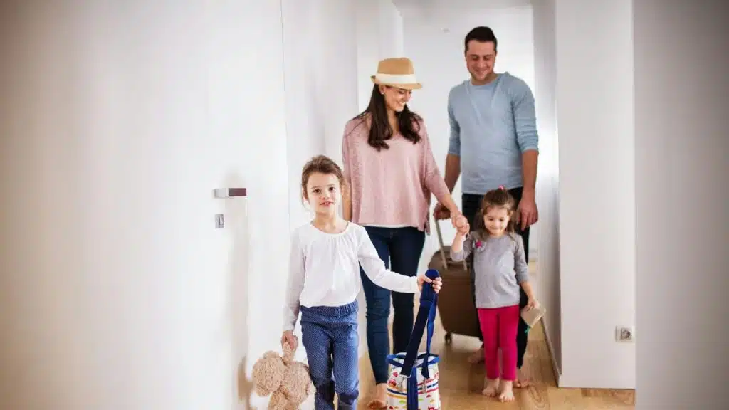 A smiling family with two young daughters and luggage is leaving their home.