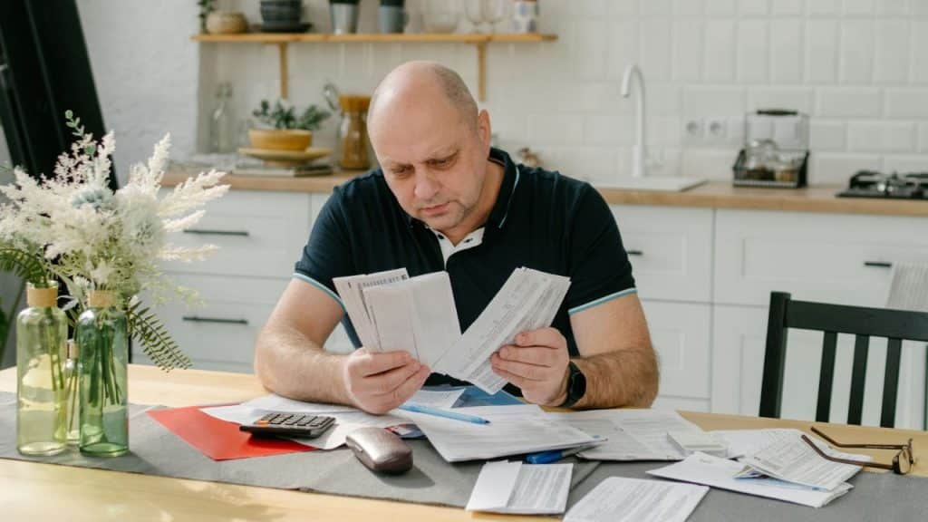 A balding man sits at a table, looking stressed while sorting through bills.