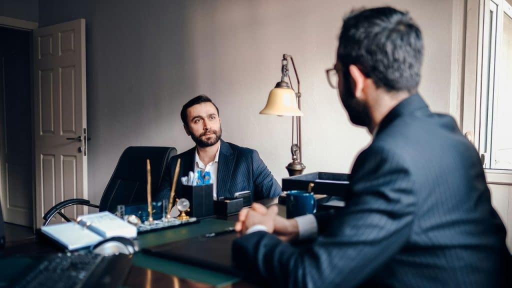 A bearded lawyer in a suit sits at his desk listening to a client.