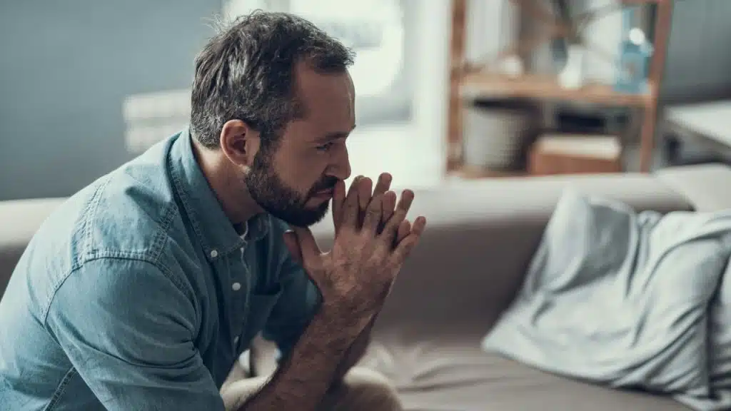 A worried, bearded man sits on a couch, staring into space with his hands clasped.
