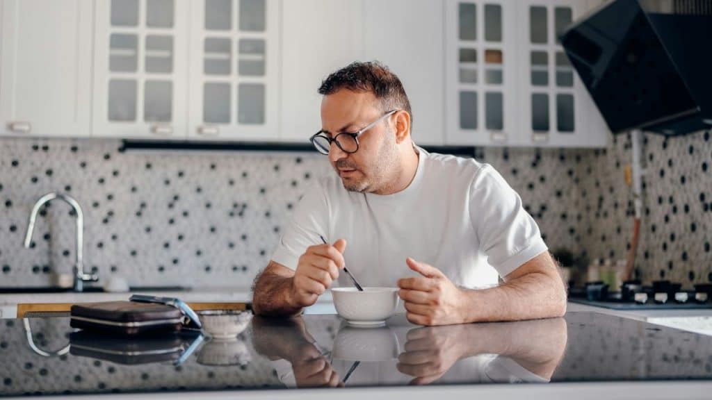 A lonely man with glasses eats cereal at a kitchen counter.