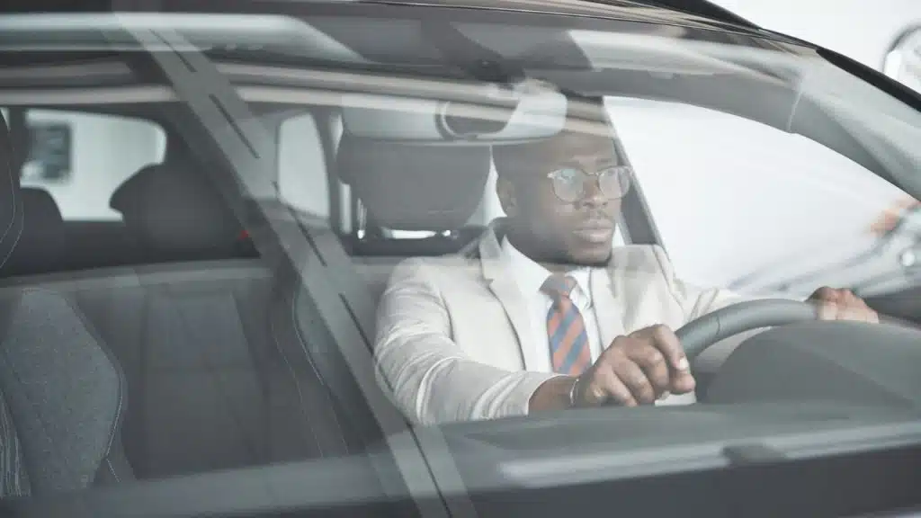 A stylishly dressed businessman with glasses and a tie sits in his car.