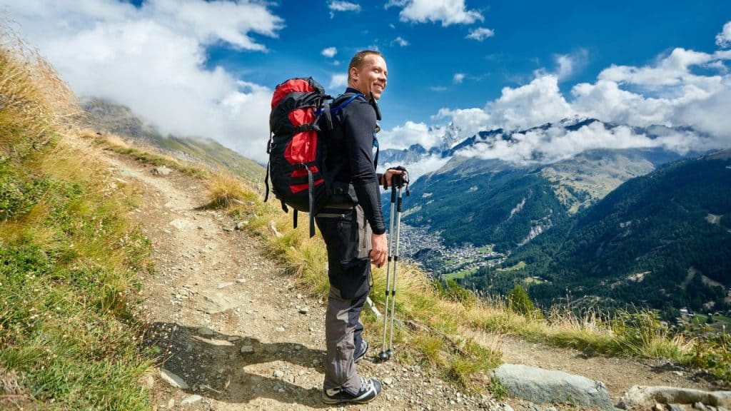 A male hiker with a backpack and poles smiles while looking out at a mountain range.