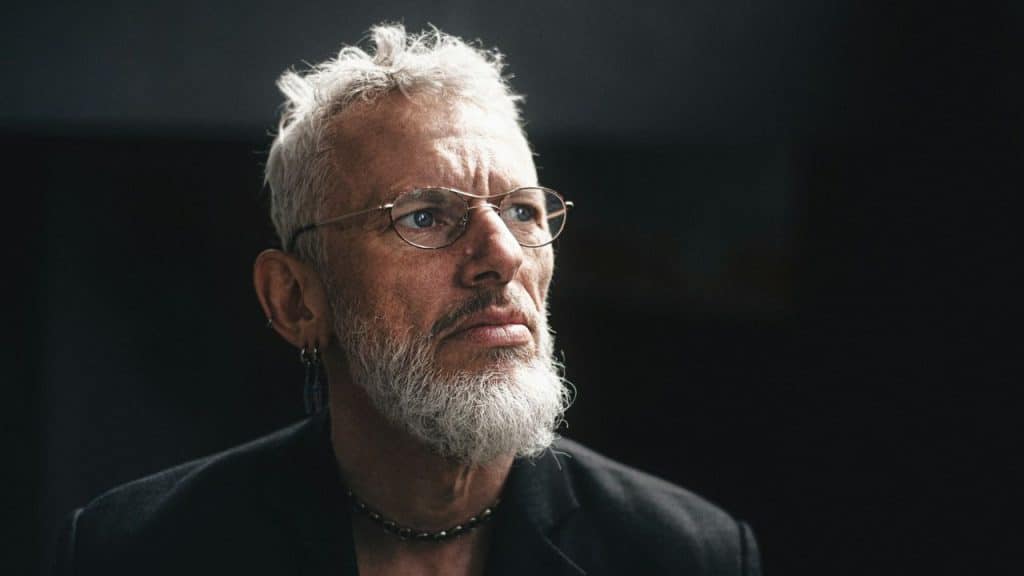 A gray-bearded man with spiked hair and glasses looks off to the side.