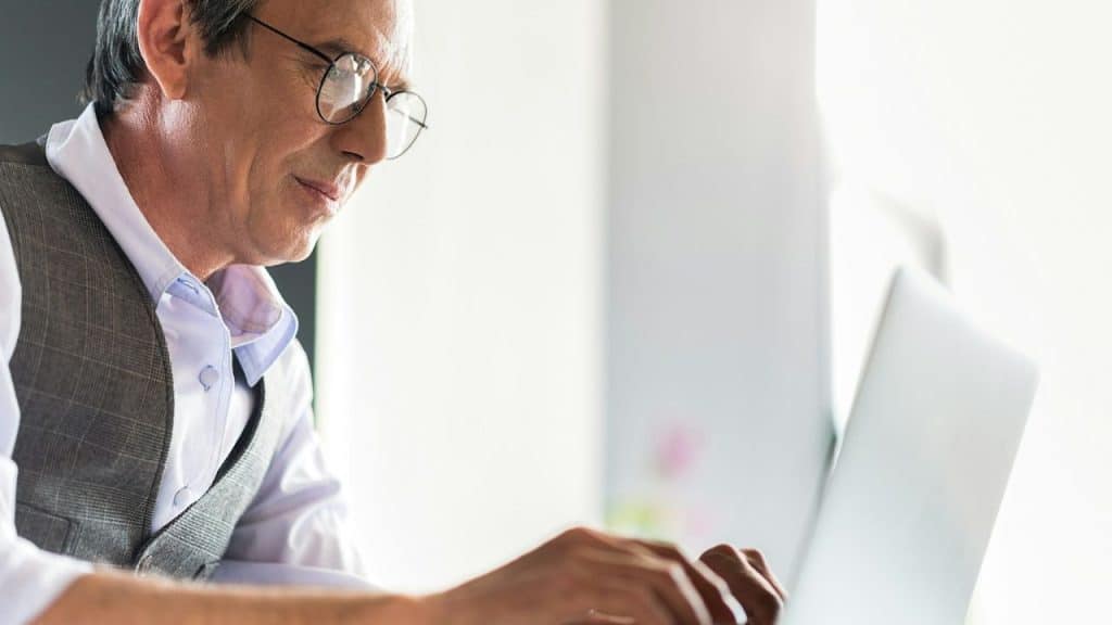 A smiling man in glasses and a vest looks down at a laptop.