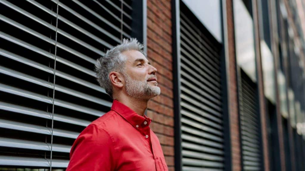 A mature man in a red shirt with gray hair and a beard, looking up and to the side.