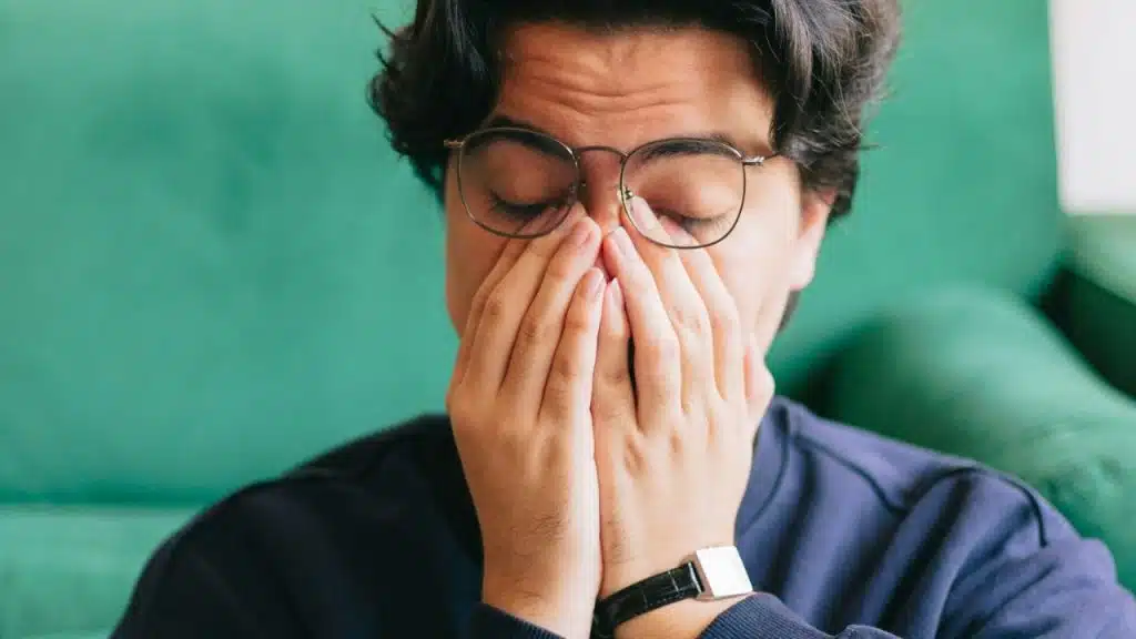 A person with glasses sits with their eyes closed, rubbing their face.