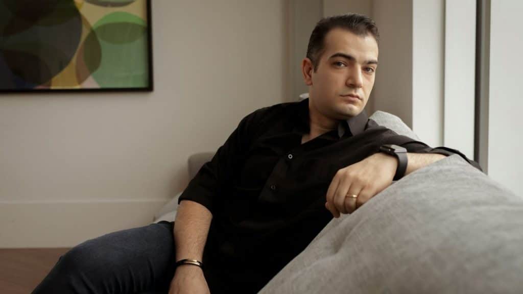 A handsome man with dark hair, a black shirt, and a gold ring sits on a gray sofa.