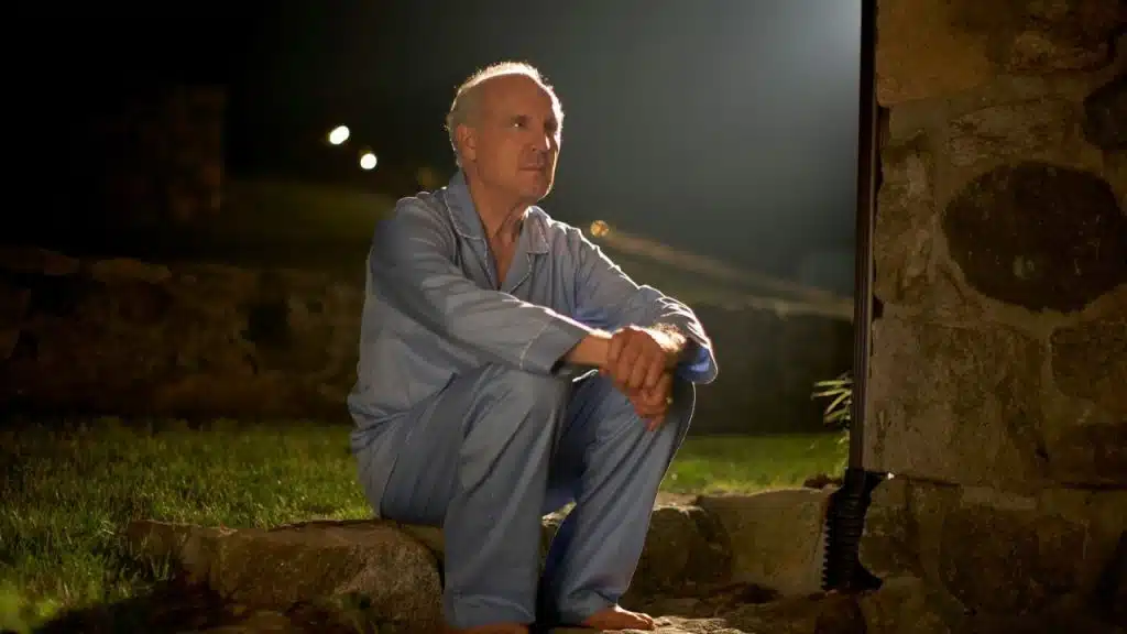 A middle-aged man in blue pajamas sits on stone steps at night.