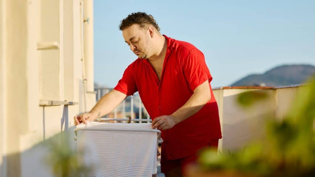 A man in a red shirt hangs up clothes on a drying rack on a balcony.
