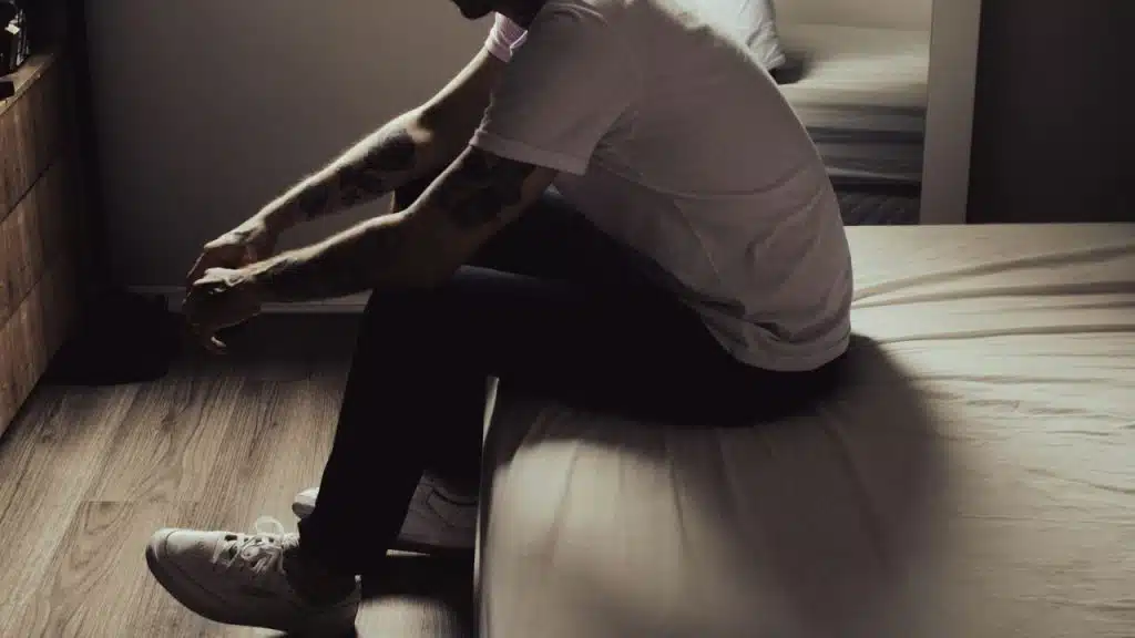 A man with tattooed arms sits on the edge of a bed, looking down.