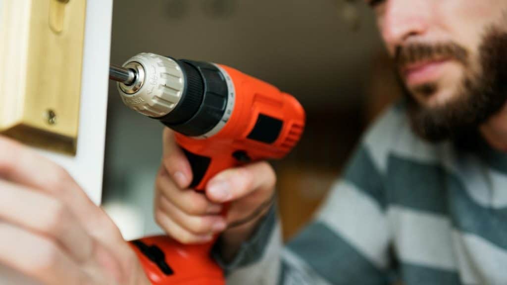 A man with a beard uses a cordless drill to fix a doorknob.