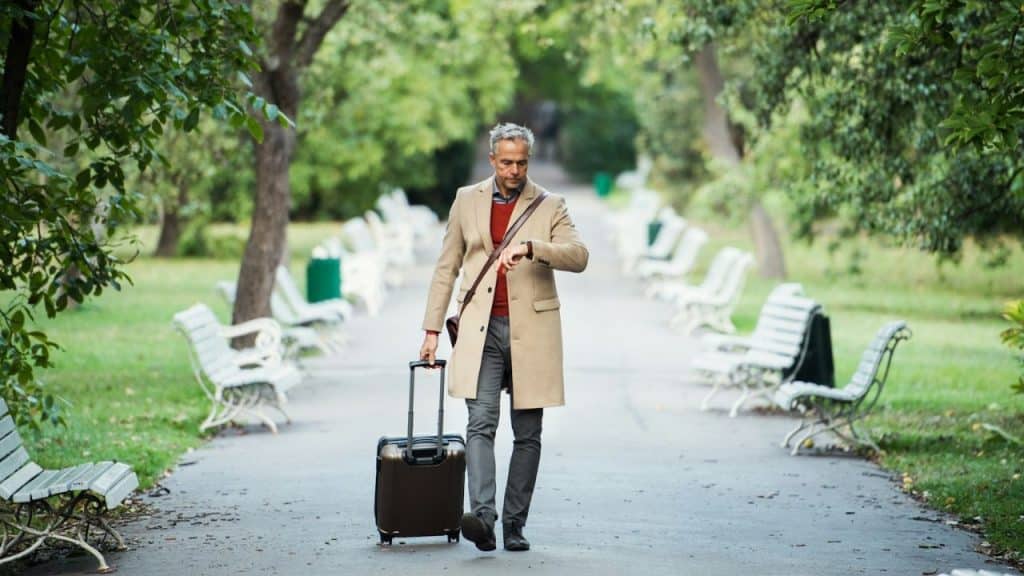 A well-dressed man with a suitcase checks his watch while walking in a park.