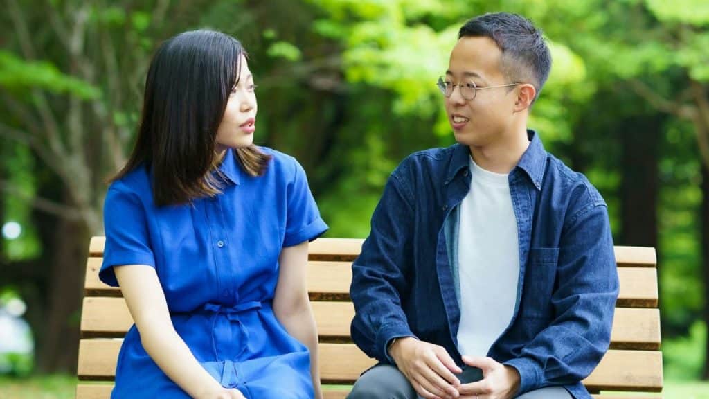 A mature couple sits on a park bench, looking at each other.