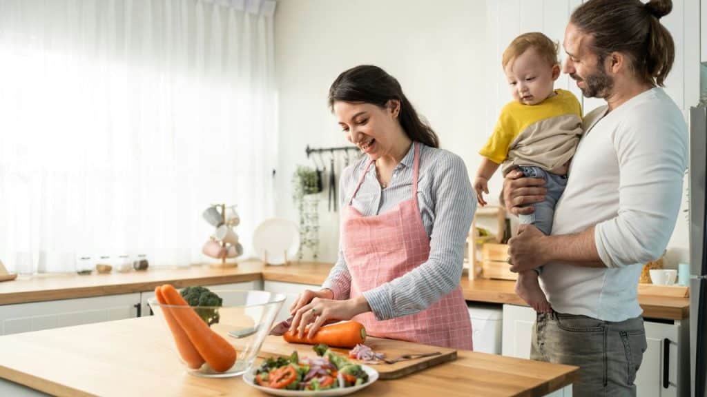 A happy family is in a kitchen, with a mother cutting carrots while a father holds a baby.