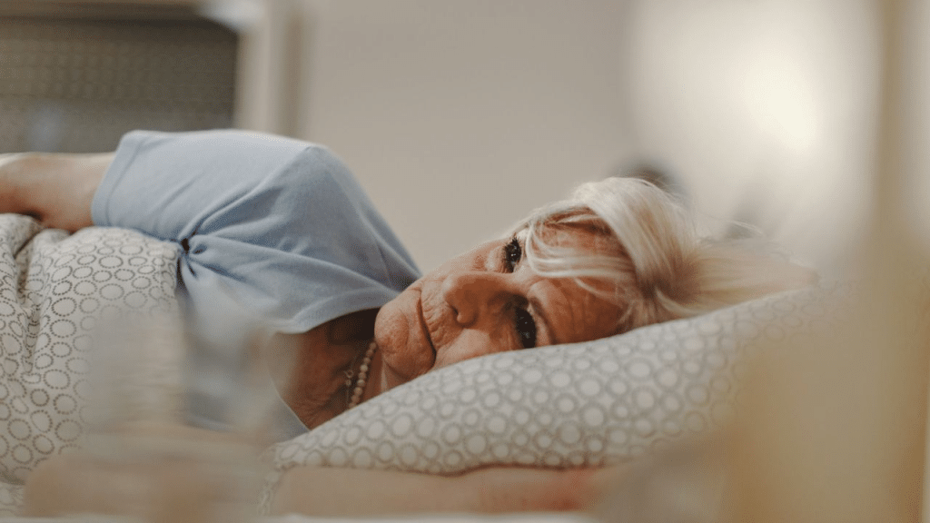 A mature woman with white hair lies in bed, propped up on a pillow. She is wearing a light blue shirt and a pearl necklace.