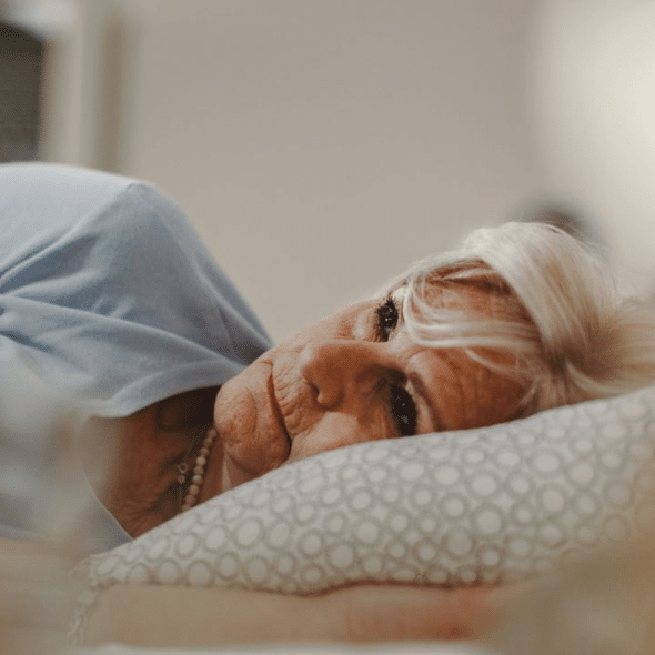 A mature woman with white hair lies in bed, propped up on a pillow. She is wearing a light blue shirt and a pearl necklace.