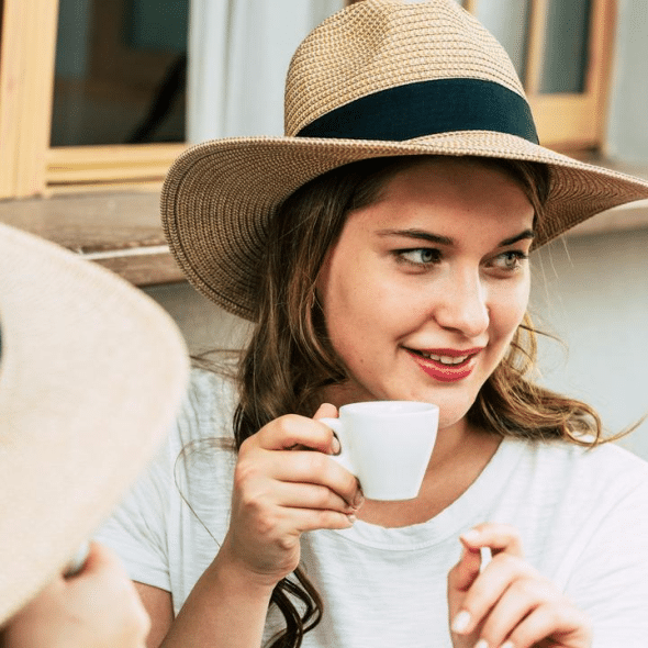 A woman wearing a straw hat smiles while holding a small cup of coffee.