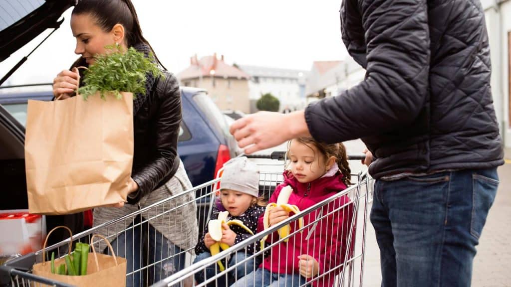 A family loading groceries into a car while children sit in a shopping cart eating bananas.
