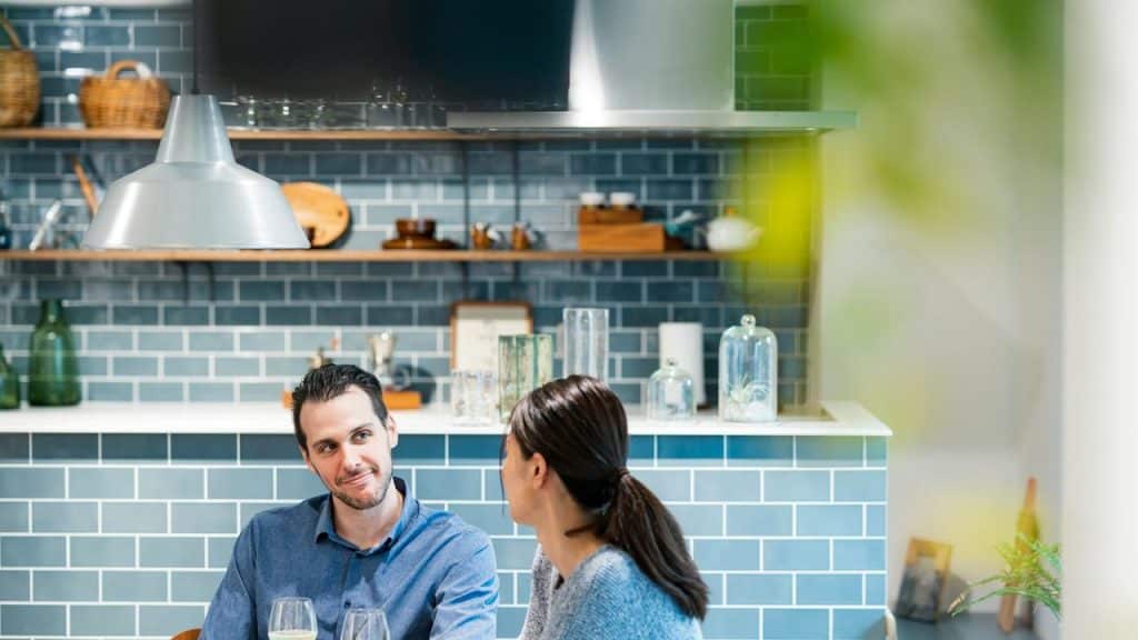 A man and woman sitting at a table in a modern kitchen with blue tile walls.
