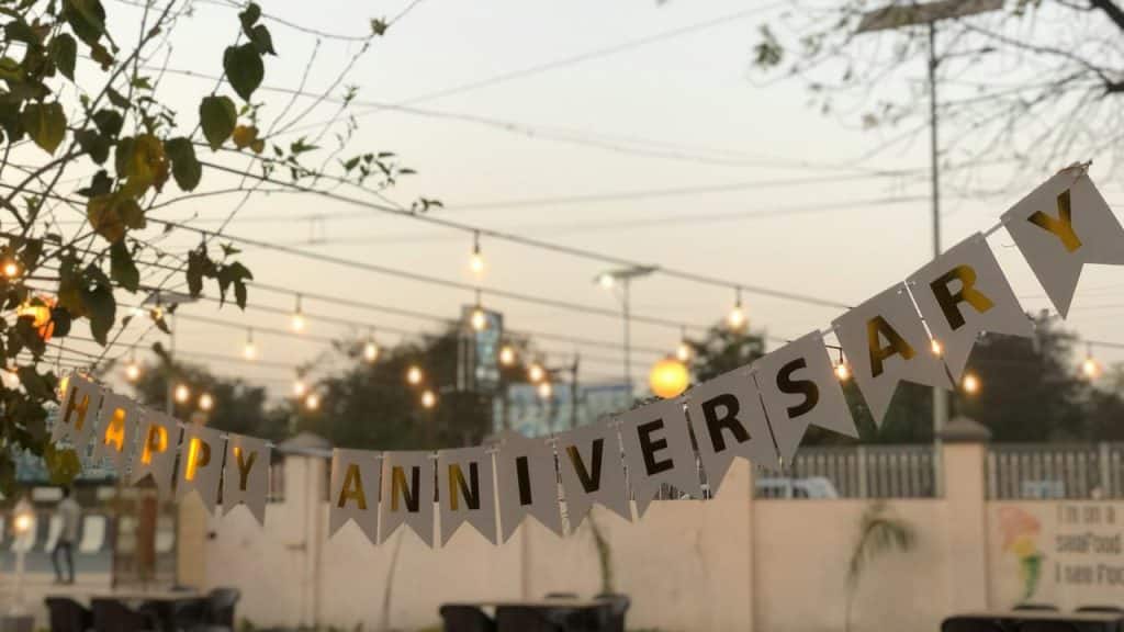A banner with "Happy Anniversary" hanging outdoors with string lights in the background.