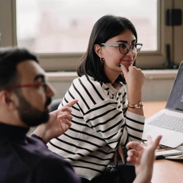 A woman in glasses listens to a colleague at a desk with a laptop.