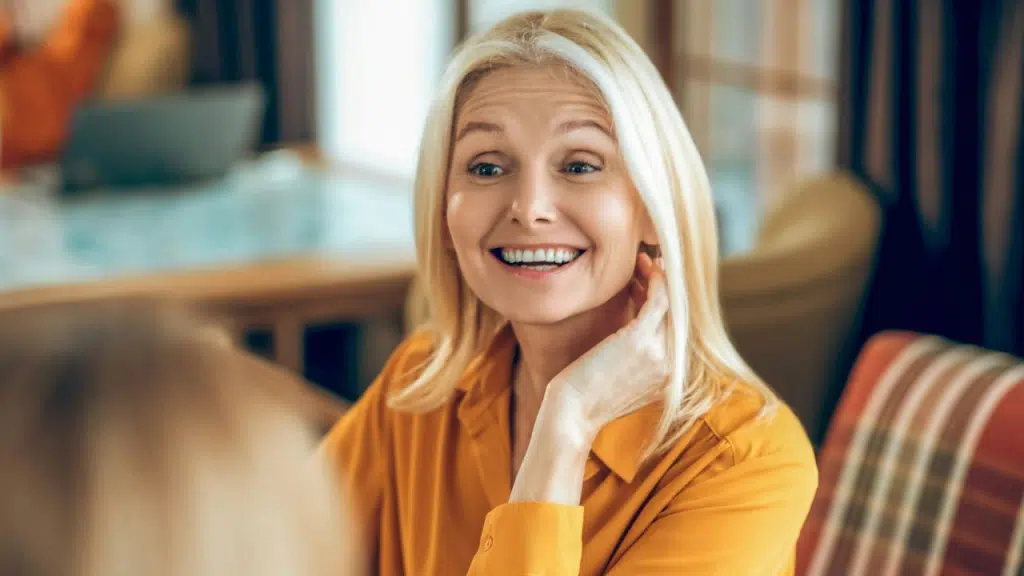 A mature blonde woman in a yellow shirt smiling and touching her hair.