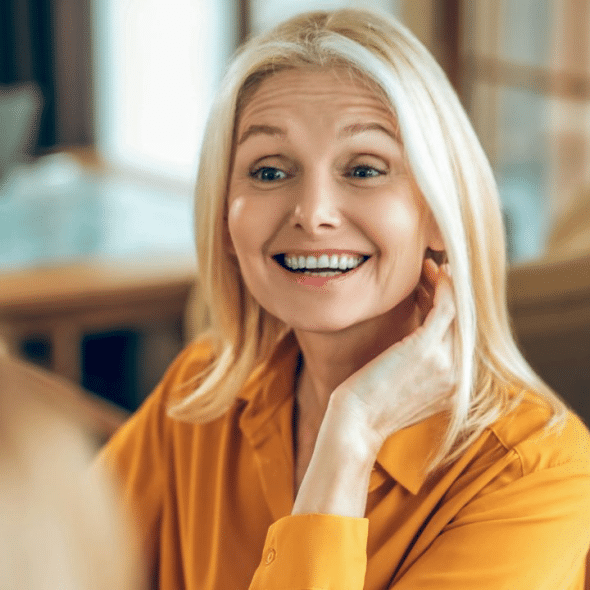 A mature blonde woman in a yellow shirt smiling and touching her hair.