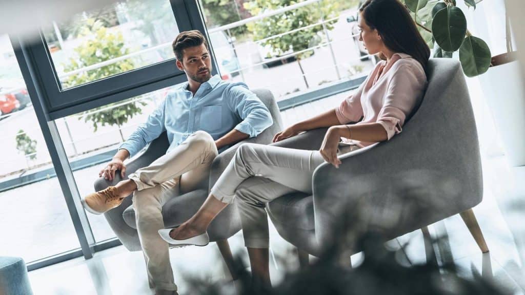 A man and woman sit in chairs having a serious conversation by a window.