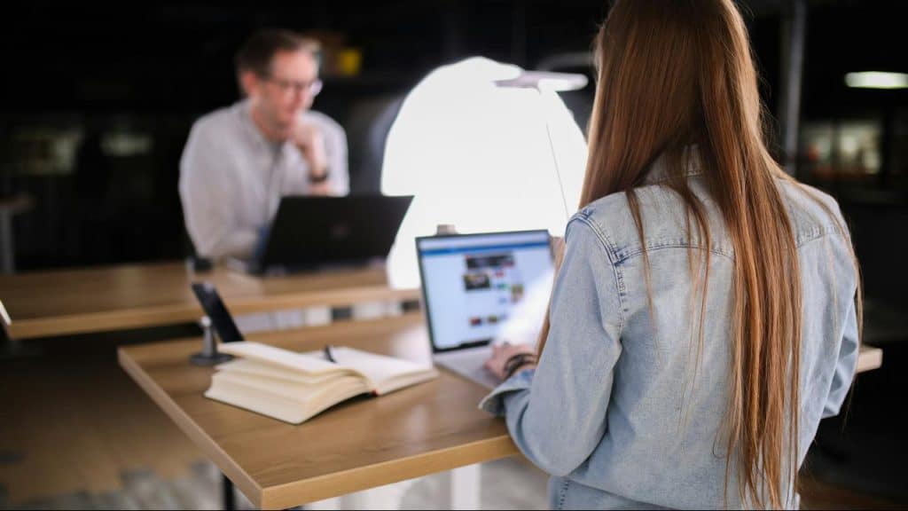 A woman works on a laptop at a standing desk with a man in the background.