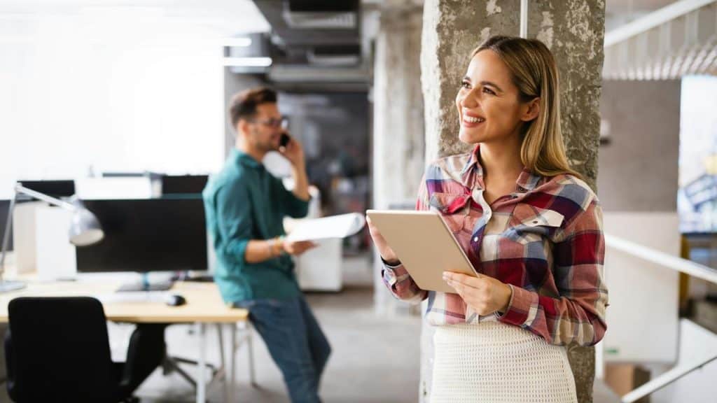 A woman smiles while holding a tablet in an office.