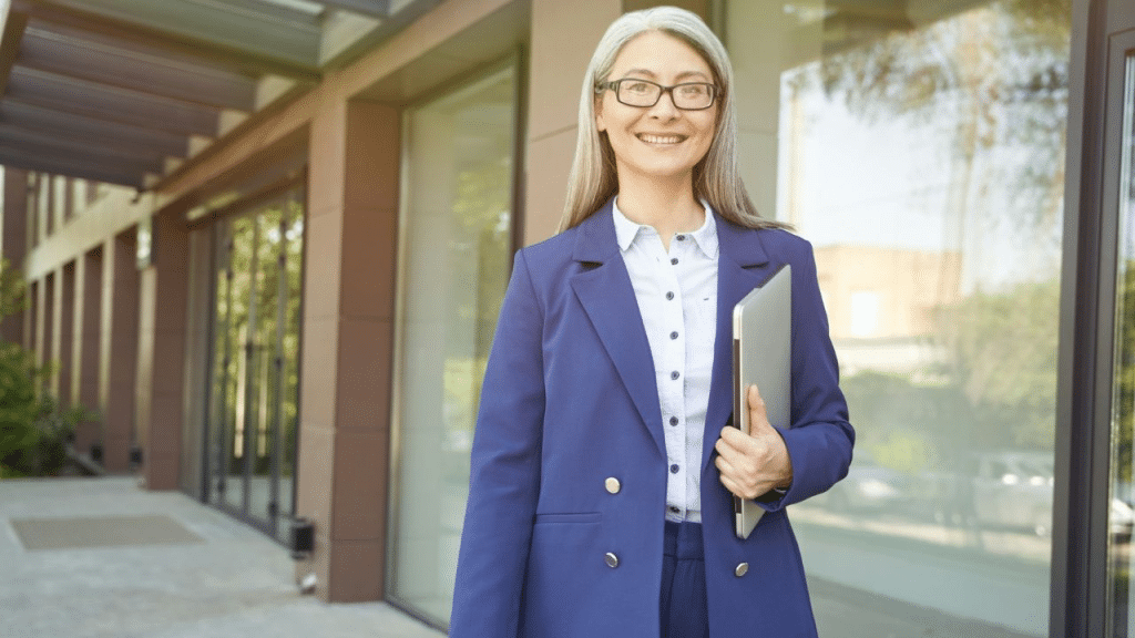 A smiling, mature businesswoman in a blue suit holding a laptop outside an office building.