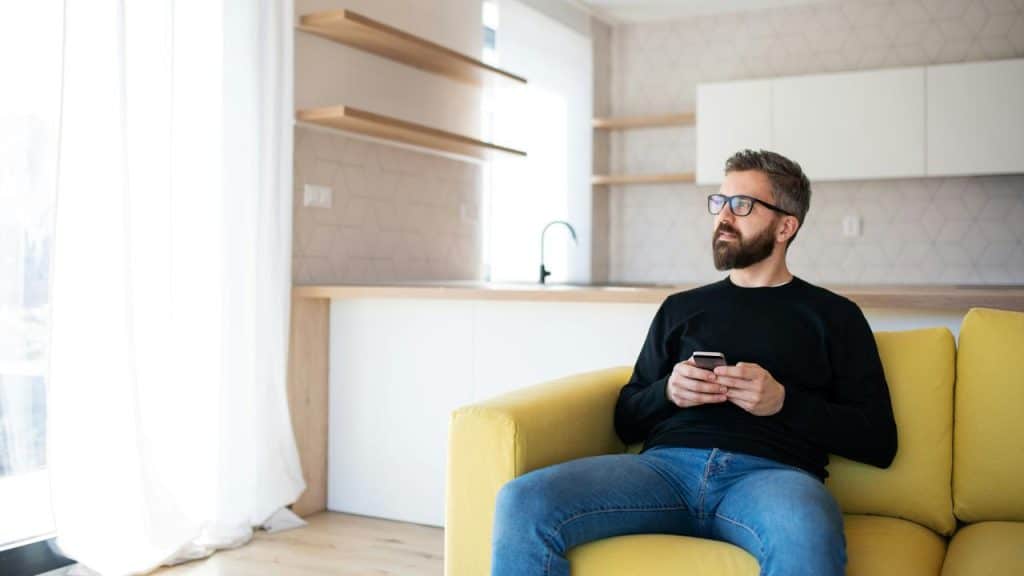 A man sitting on a yellow couch holding a smartphone and looking out the window.