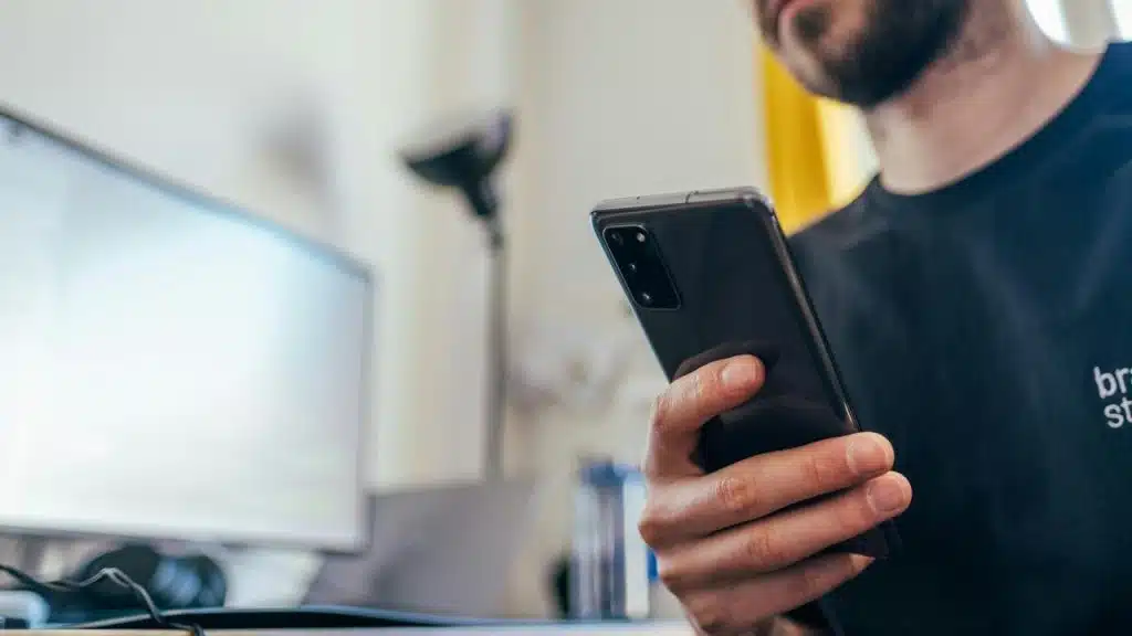 A man holding a smartphone while sitting at a desk with a computer.