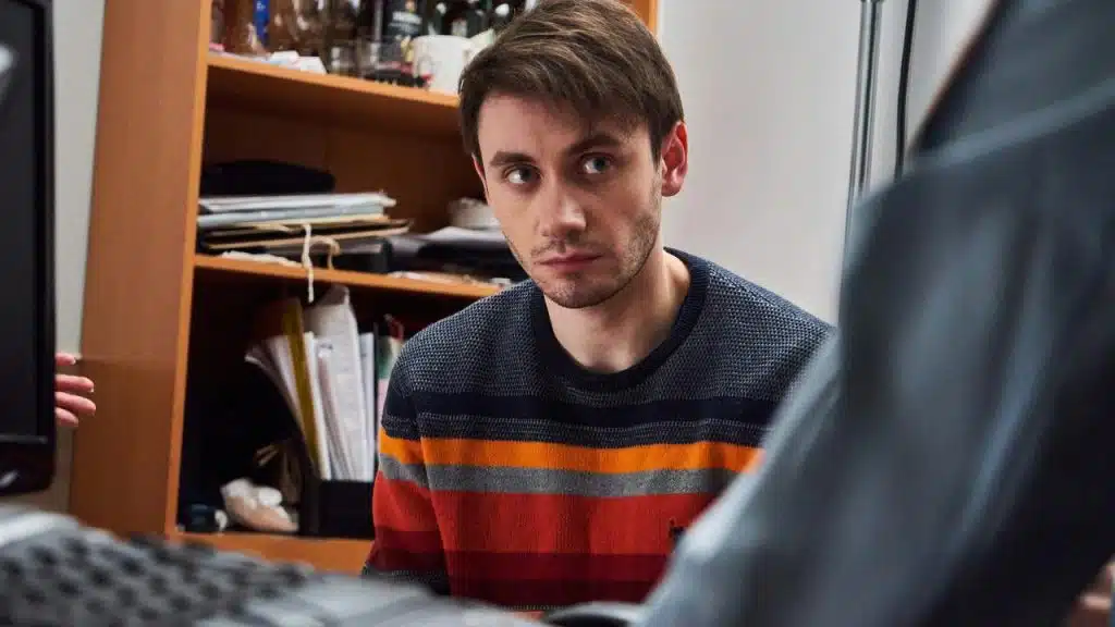 A man in a striped sweater sitting indoors with a serious expression.