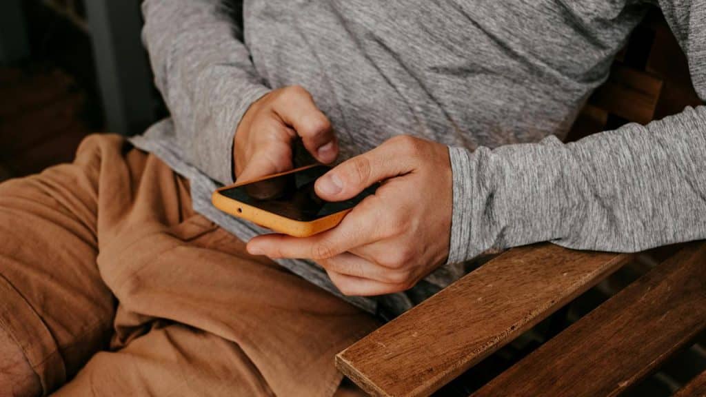 A person holding a smartphone with an orange case while sitting on a wooden chair.