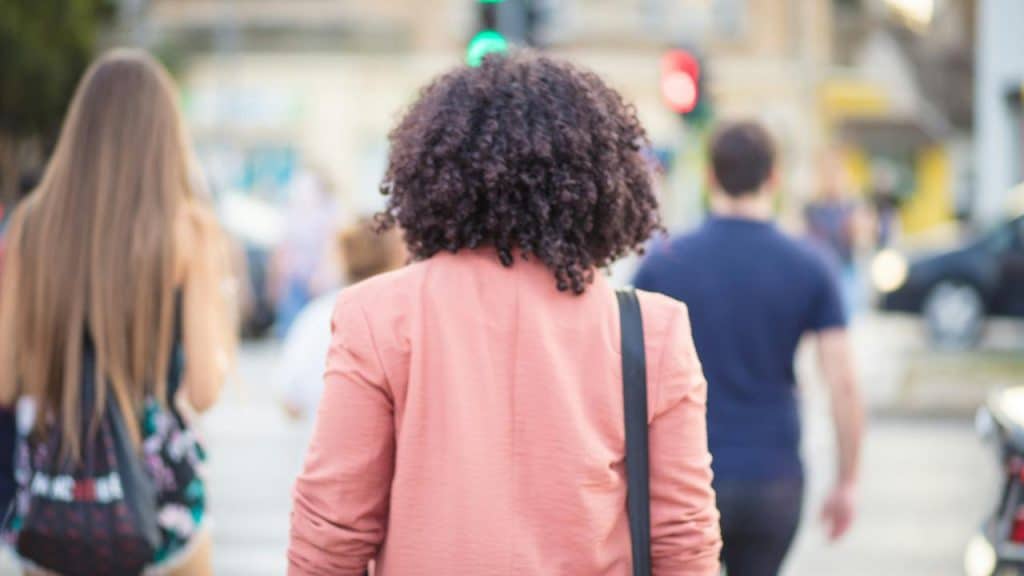 A person with curly hair wearing a pink jacket walking on a city street.