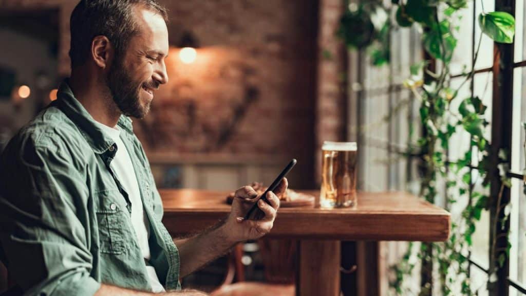 A man smiling while looking at his phone with a glass of beer on the table.