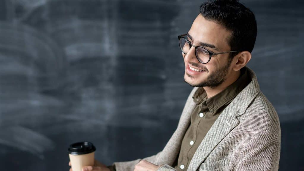 A man wearing glasses and a blazer smiling while holding a coffee cup.