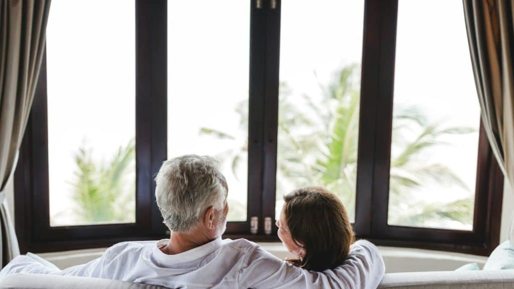 An older couple sitting together on a couch, looking out a large window at palm trees.