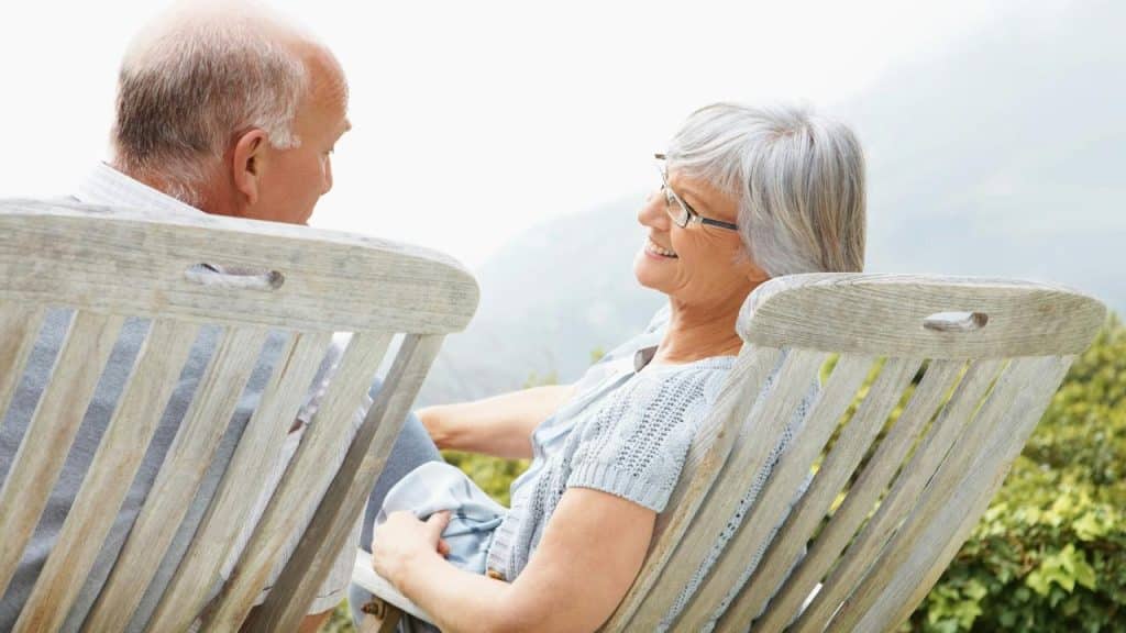 An elderly couple sitting on wooden chairs outdoors, smiling and talking to each other.