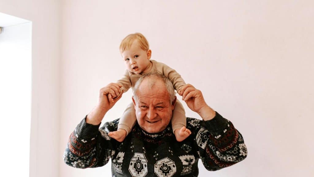 An elderly man smiling while carrying a baby on his shoulders.