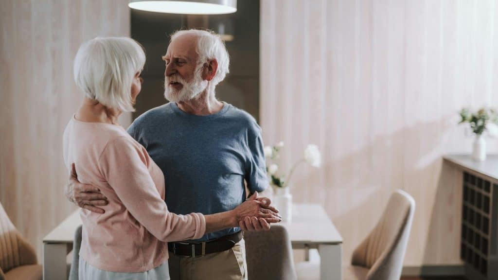 An elderly couple dancing together in their dining room while smiling at each other.