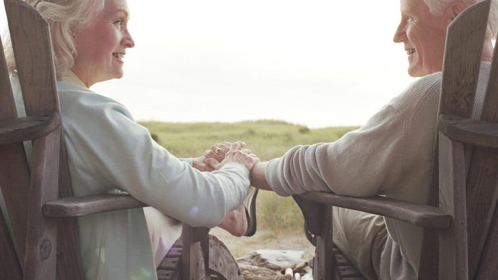An elderly couple sitting outdoors in wooden chairs and smiling at each other.