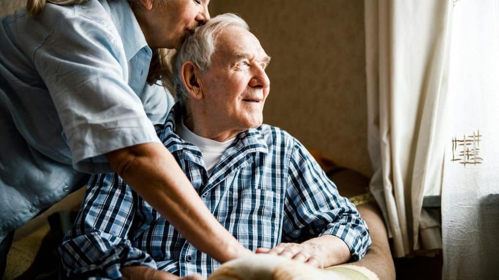 An elderly woman gently kissing the forehead of an elderly man while holding his shoulder.