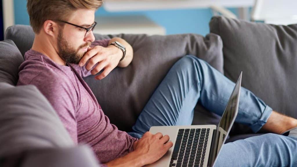 A man sitting on a couch using a laptop while looking thoughtful.