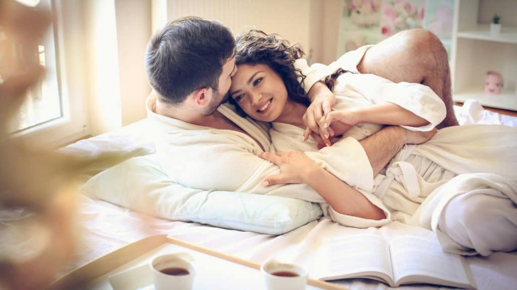 A couple in white robes cuddling in bed while smiling at each other.